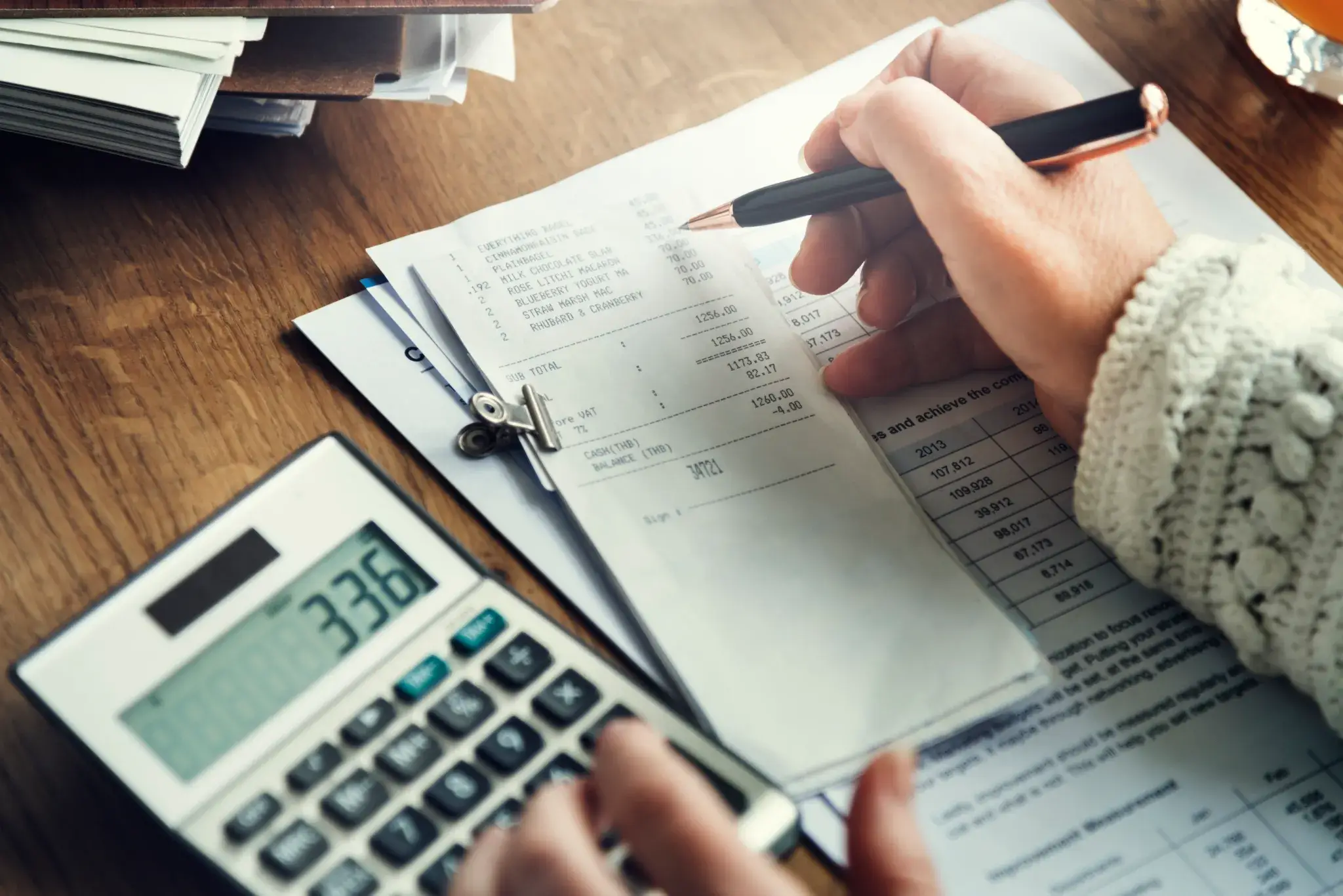 A person is calculating expenses using a calculator while reviewing receipts and financial documents on a wooden desk.