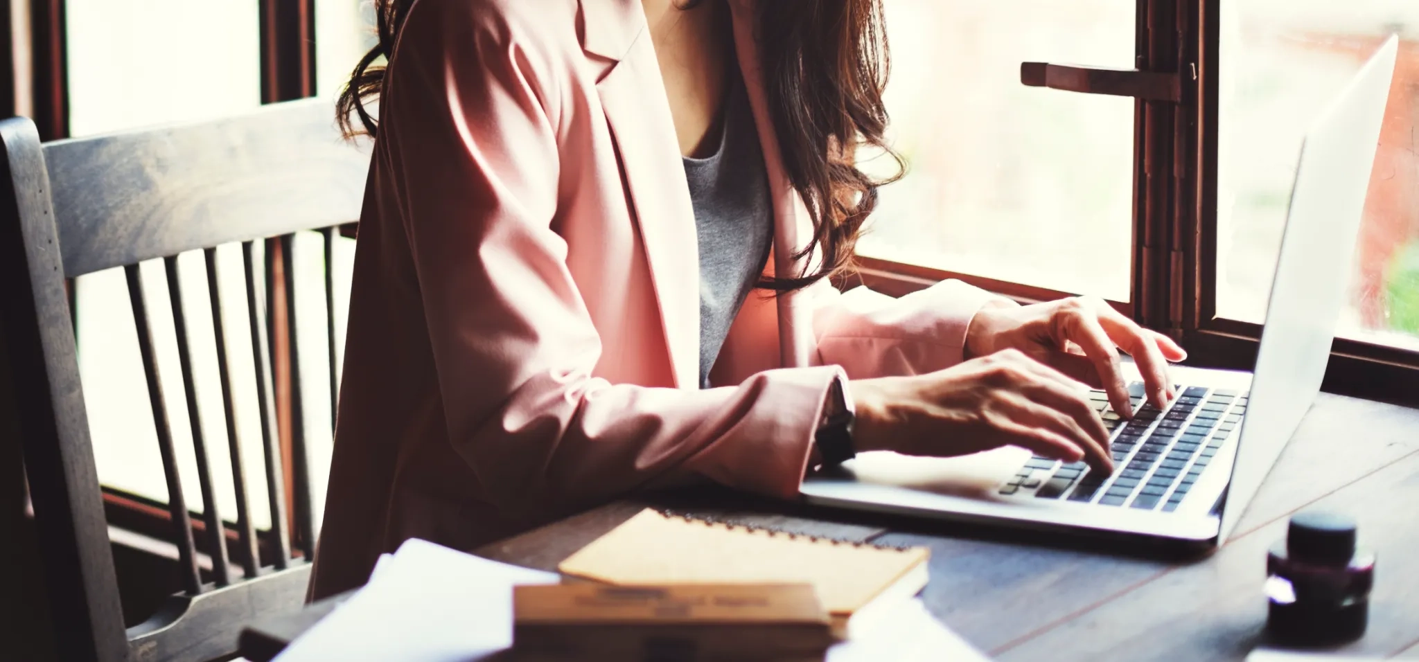 A female corporate secretary in pink blazer typing on a laptop by a window, working at a wooden desk with notebooks, papers, and an ink bottle nearby